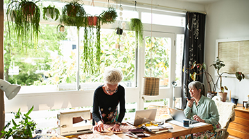 Two older women working in a sunlit craft room filled with plants; one uses a sewing machine while the other types on a laptop at a shared wooden table.
