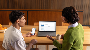 Young adult and a woman sitting at a table discussing information on a smartphone and laptop, with a mug and bowl of oranges nearby.
