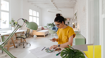 Designer in a bright studio holding a smartphone and color swatches, reviewing plans on a desk with papers and plants.